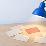 Lampe d'architecte bleue dont le faisceau isole une fiche corail parmi des fiches crème posées sur un bureau clair : le cadrage qu'apporte un consultant chatbot IA